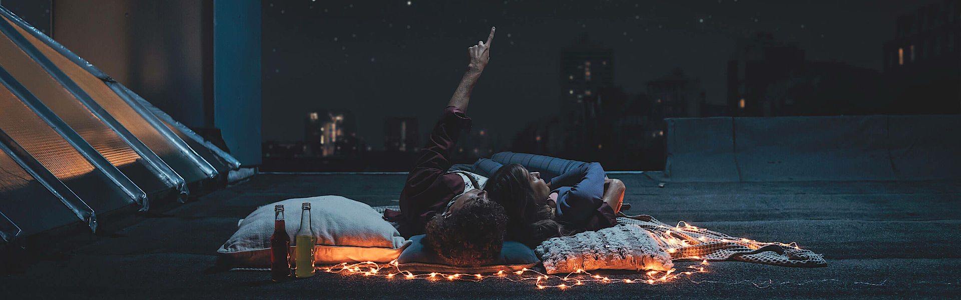 Ein Paar liegt auf der Dachterrasse und betrachtet den Sternenhimmel. Der Mann zeigt mit dem Arm in den Himmel.