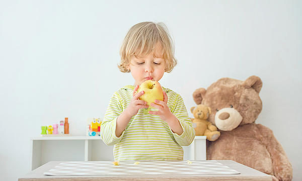 Kleinkind steht hinter einem Spieltisch und isst einen Apfel. Ein großer Teddybär sitzt im Hintergrund.