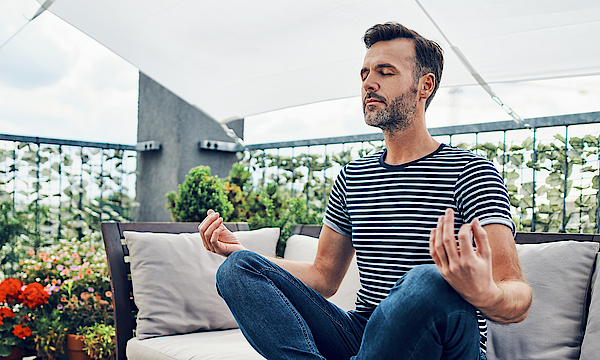 Mann ist draußen auf dem Balkon und sitzt im Schneidersitz in einer Entspannungsposition auf der Bank.