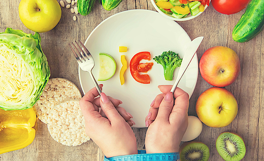 Auf einem Tsich liegt verschiedenes Gemüse. Auf einem weißen Teller ist daraus das Wort Diät geschrieben. Hände halten Besteck.