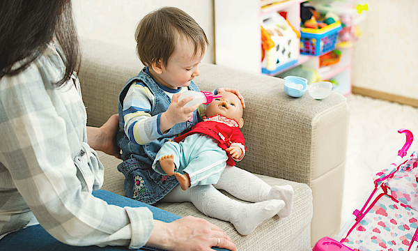 Eine Mutter sitzt mit ihrer Tochter auf dem Sofa. Das Mädchen füttert eine Babypuppe mit der Flasche.