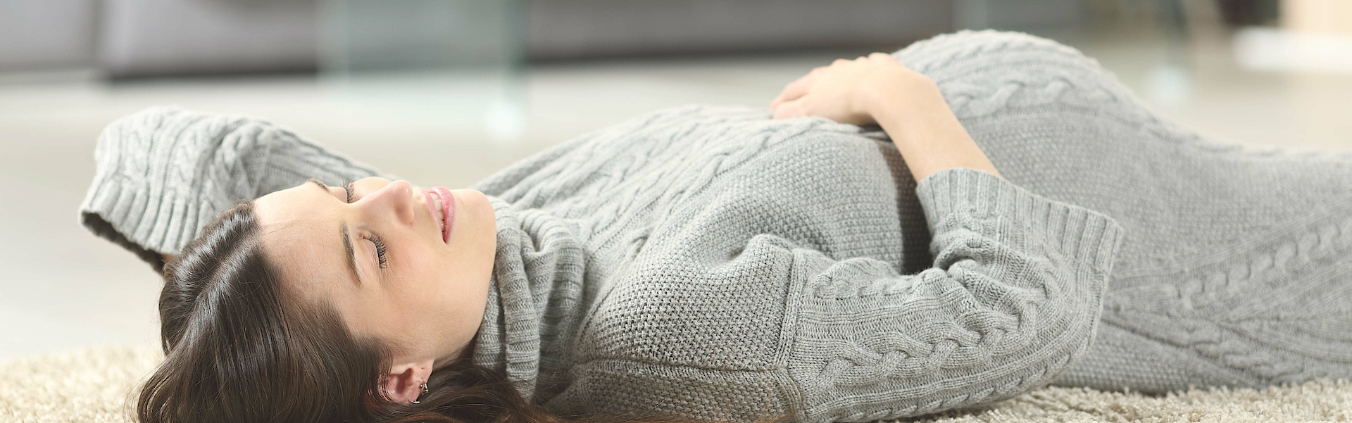Eine schwangere junge Frau liegt auf einem Teppich. Sie hat ein graues Strickkleid an. Ihre rechte Hand ruht entspannt auf ihrem Bauch. Sie hat die Augen geschlossen und entspannt sich.