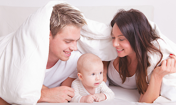 Ein Elternpaar liegt zugedeckt mit dem Baby im Bett.