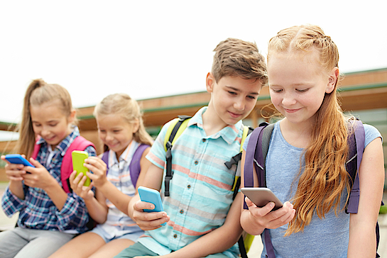 Drei Mädchen und ein Junge sitzen beisammen. Sie alle haben ein Smartphone in der Hand. Der Junge schaut mit auf auf das Smartphone des Mädchens neben ihm. Die anderen beiden Mädchen schaunen auf ihre Displays.