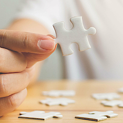 Eine Frau sitzt an einem Tisch. Sie legt ein Puzzle zusammen. Ein Teil hält sie in der Hand.