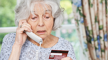 Eine ältere Frau mit grauen Haaren telefoniert. Sie macht ein besorgtes Gesicht. In der Hand hält sie ihre Bankkarte. Sie gibt dem Anrufer ihre Bankdaten durch.