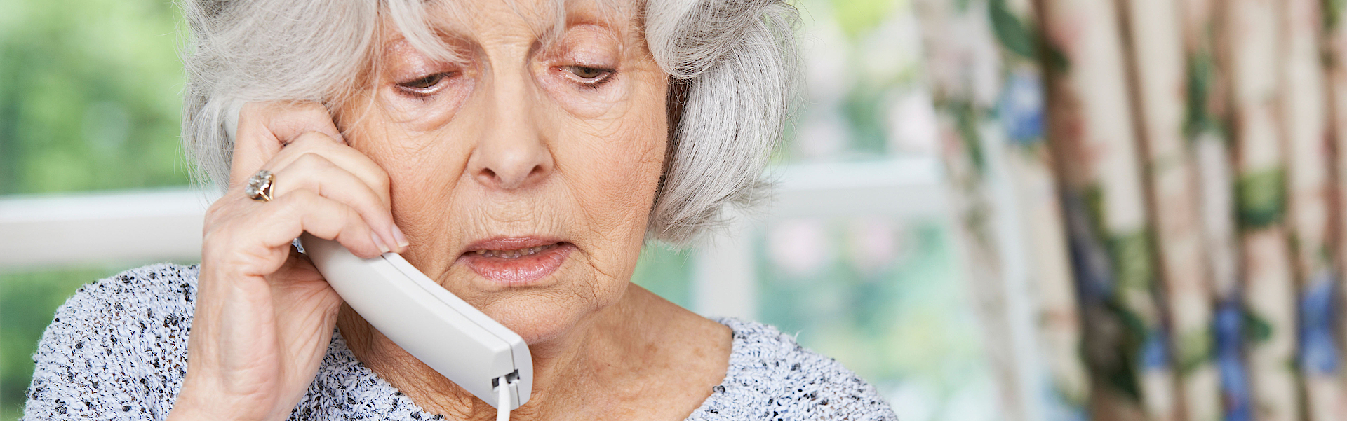 Eine ältere Frau mit grauen Haaren telefoniert. Sie macht ein besorgtes Gesicht. In der Hand hält sie ihre Bankkarte. Sie gibt dem Anrufer ihre Bankdaten durch.