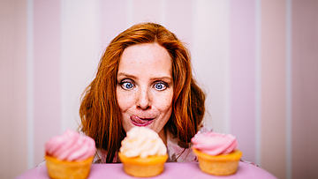 Eine junge Frau sitzt vor drei Cupcakes. Sie möchte diese wirklich essen und schaut ganz begierig. Sie sind voller Zucker und sehen sehr lecker aus. Die junge Frau traut sich aber nicht wirklihch, die Cupcakes zu essen. Ihr läuft das Wasser im Mund zusammen.
