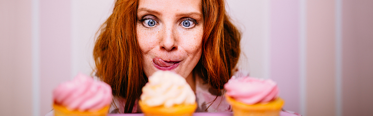 Eine junge Frau sitzt vor drei Cupcakes. Sie möchte diese wirklich essen und schaut ganz begierig. Sie sind voller Zucker und sehen sehr lecker aus. Die junge Frau traut sich aber nicht wirklihch, die Cupcakes zu essen. Ihr läuft das Wasser im Mund zusammen.