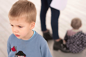 Ein kleiner Junge steht abseits seiner Mutter und des Geschwisterchens. Er ist eifersüchtig auf das Geschwisterchen und macht ein enttäuschtes Gesicht.