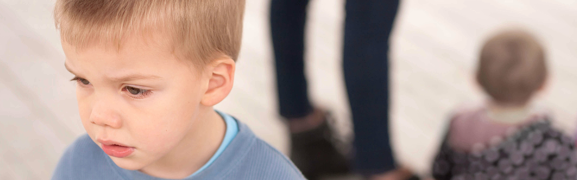 Ein kleiner Junge steht abseits seiner Mutter und des Geschwisterchens. Er ist eifersüchtig auf das Geschwisterchen und macht ein enttäuschtes Gesicht.