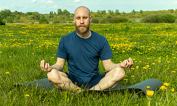 Ein Mann in sportlicher Kleidung sitzt auf einer Blumenwiese. Um ihn herum blüht Löwenzahn. Er meditiert im Schneidersitz auf einer Yogamatte.