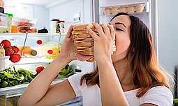 Eine Frau steht vor dem geöffneten Kühlschrank und isst ein dickes Käse-Schinken Sandwich. Der Kühlschrank ist gefüllt mit Lebensmitteln.
