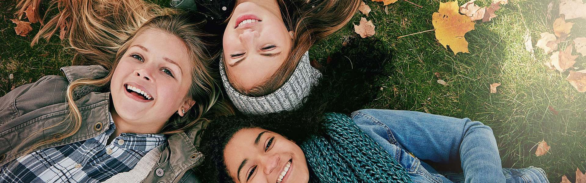 Drei junge Mädchen mit schönen Zähnen liegen auf einer Herbstwiese und lachen in die Kamara.