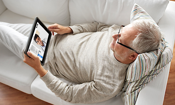 Ein älterer Mann liegt auf einem Sofa. Er hat ein Tablet in der Hand. Dort ist sein Arzt zu sehen, der ihn berät.