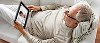 Ein älterer Mann liegt auf einem Sofa. Er hat ein Tablet in der Hand. Dort ist sein Arzt zu sehen, der ihn berät.