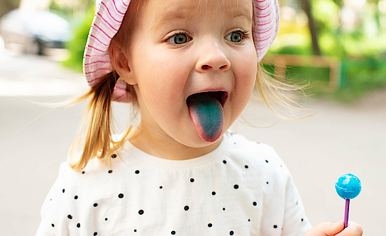 Kind steckt blaue Zunge raus und hält einen blauen Lolli in der Hand.