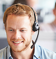 In einem Callcenter sitzt ein junger, blonder Mann mit Jeanshemd. Er hat ein Headset auf und telefoniert.