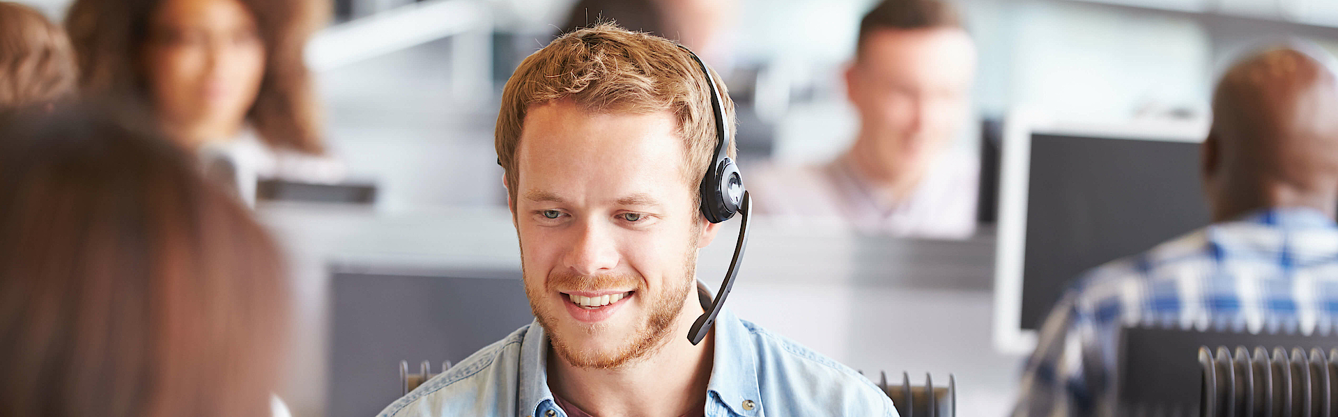 In einem Callcenter sitzt ein junger, blonder Mann mit Jeanshemd. Er hat ein Headset auf und telefoniert.