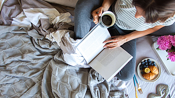 Frau sitzt mit einer Tasse Kaffee und einem Buch auf dem Bett