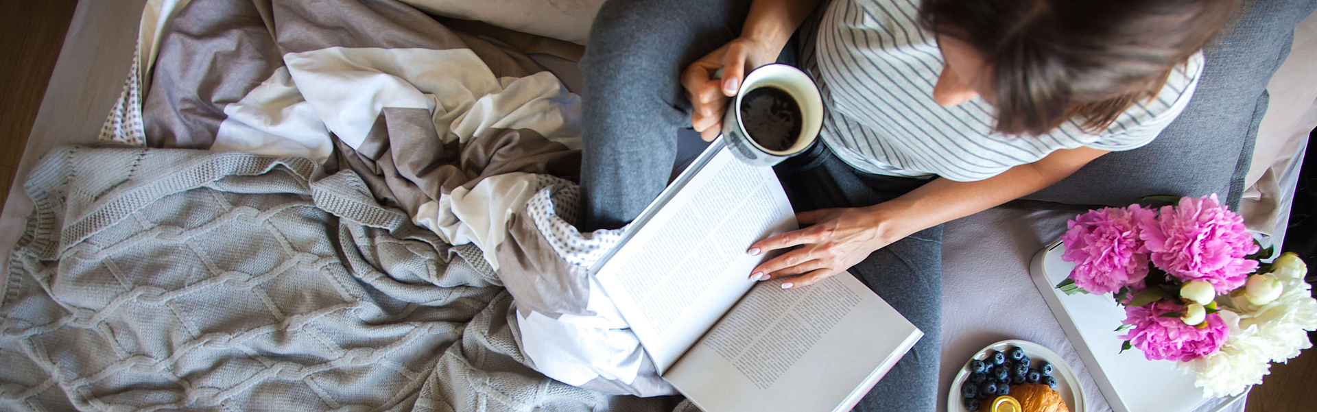 Frau sitzt mit einer Tasse Kaffee und einem Buch auf dem Bett