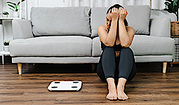 Eine asiatische Frau sitzt vor einem grauen Sofa neben einer Waage und betrachtet diese. Sie ist unglücklich.