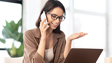 Fröhliche, junge Frau mit Brille telefoniert und schaut dabei in einen Laptop.
