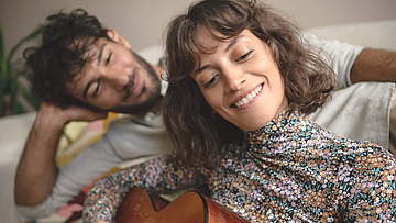 Junges Paar glückliches liegt auf einem Bett, sie spielt Gitarre und er schaut ihr dabei zu.