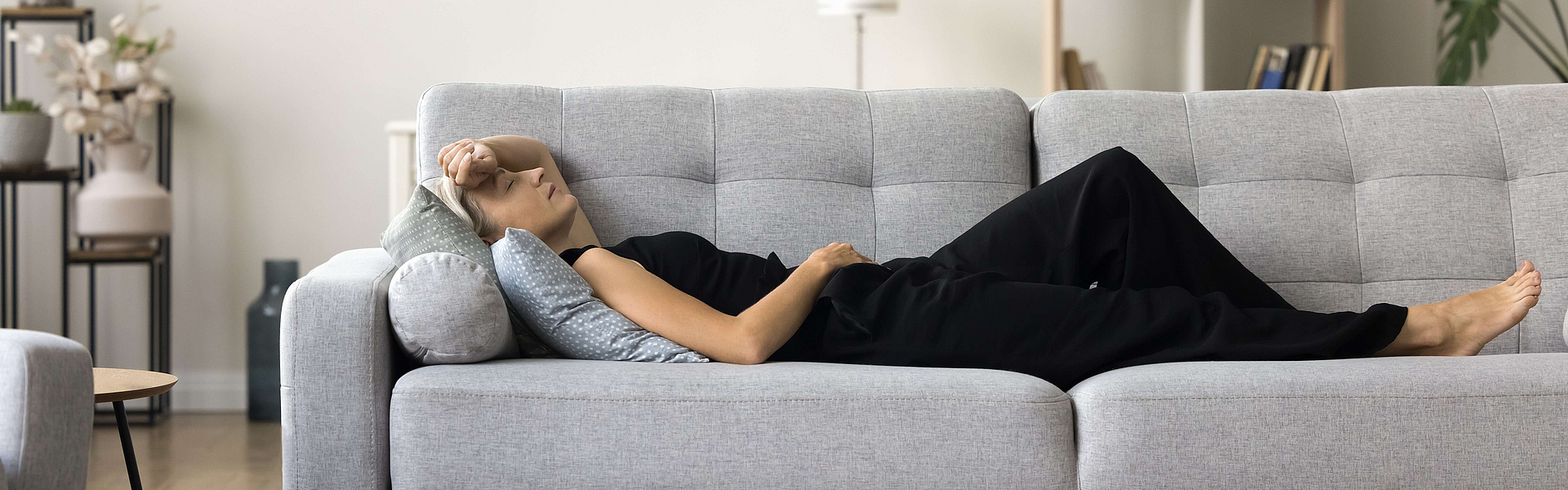 Eine müde Frau liegt auf einem grauen Sofa und ruht sich aus.