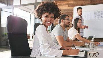 Lächelnde Mitarbeitende sitzen an einem Konferenztisch, ein Mann arbeitet am Whiteboard.