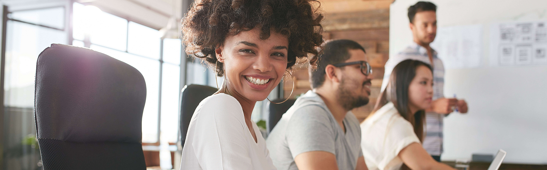 Lächelnde Mitarbeitende sitzen an einem Konferenztisch, ein Mann arbeitet am Whiteboard.