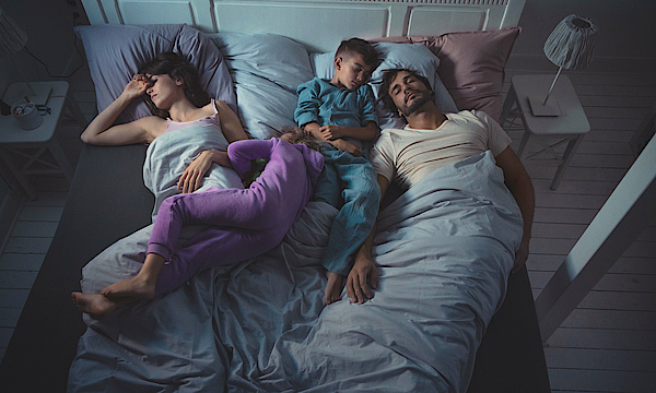 Eine Familie liegt im Bett und schläft. Außen die Eltern, innen liegen die Kinder zwischen den Eltern.
