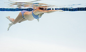 Eine schwangere Frau in einem blau-weiß gestreiften Bikini schwimmt bäuchlings in einem Schwimmbecken. Ihr rechter Arm ist nach vorn ausgestreckt, als würde sie kraulen. Sie schwimmt mit dem Gesicht unter Wasser. 