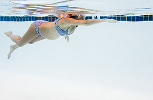 Eine schwangere Frau in einem blau-weiß gestreiften Bikini schwimmt bäuchlings in einem Schwimmbecken. Ihr rechter Arm ist nach vorn ausgestreckt, als würde sie kraulen. Sie schwimmt mit dem Gesicht unter Wasser. 