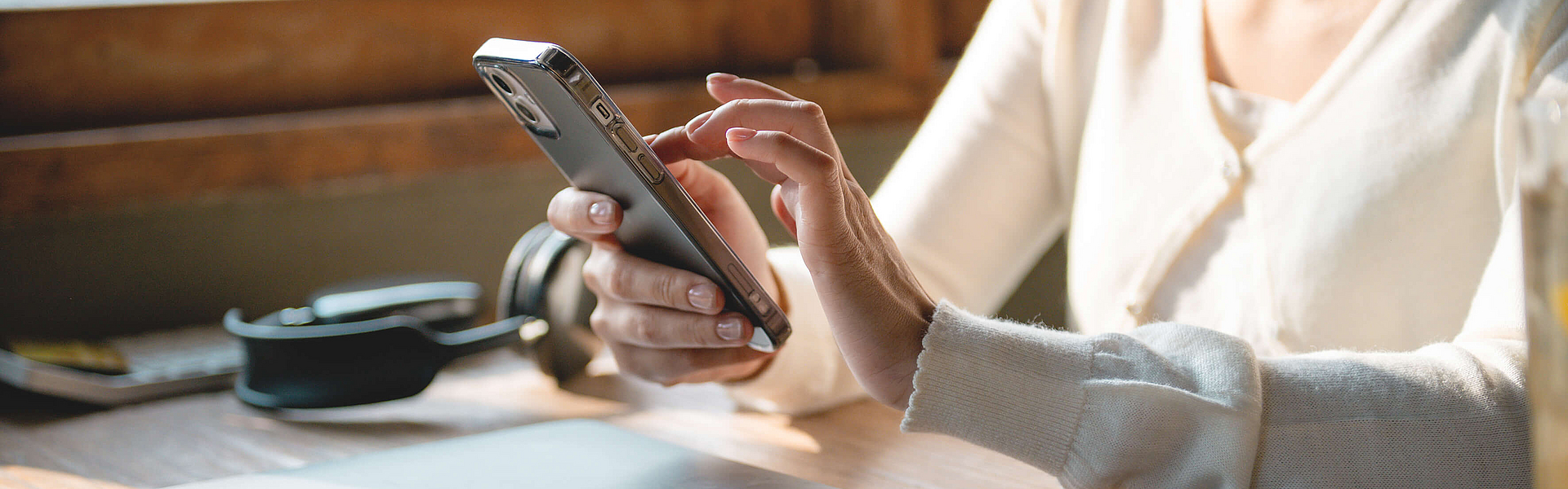 Frau tippt in das Smartphone, vor ihr auf dem Tisch liegt ein Laptop.