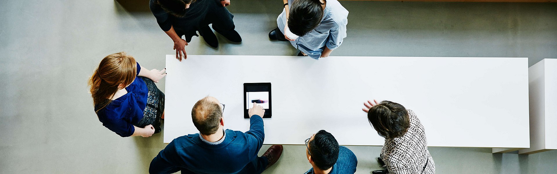 Vogelperspektive von oben auf sechs Personen in Business-Umgebung, die um einen großen weißen Tisch herumstehen und nach unten auf ein Tablet schauen.