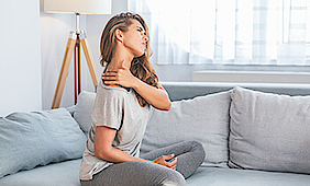 Frau sitzt auf dem Sofa. Sie berührt mit der Hand Ihre Schulter, sie hat Schmerzen.