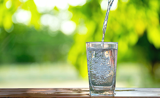 Auf einem Tisch steht ein Glas Wasser. Es wird eingeschenkt.