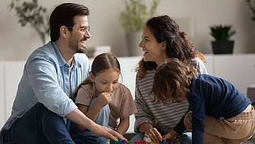 Fröhliche Eltern und zwei kleine Kinder die mit Spielzeug auf dem Teppich spielen.