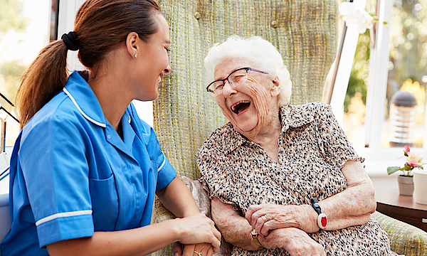 Eine Seniorin sitzt im Rollstuhl. Neben ihr eine Pflegerin im blauen Oberteil. Sie lachen beide herzlich. Ihnen geht es gut. Sie sind zusammen draußen in der Natur.