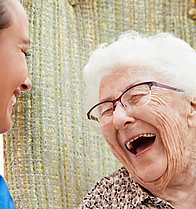 Eine Seniorin sitzt im Rollstuhl. Neben ihr eine Pflegerin im blauen Oberteil. Sie lachen beide herzlich. Ihnen geht es gut. Sie sind zusammen draußen in der Natur.