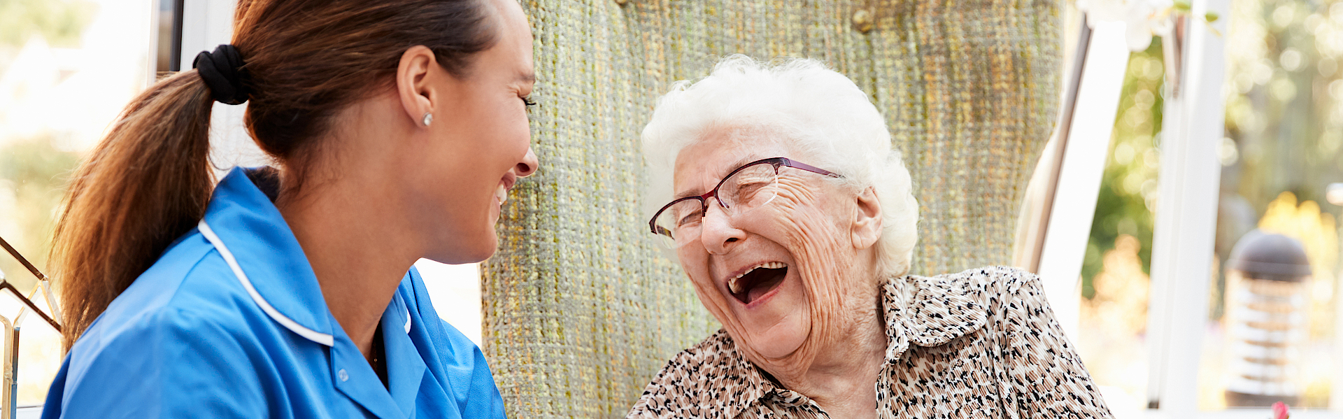 Eine Seniorin sitzt im Rollstuhl. Neben ihr eine Pflegerin im blauen Oberteil. Sie lachen beide herzlich. Ihnen geht es gut. Sie sind zusammen draußen in der Natur.