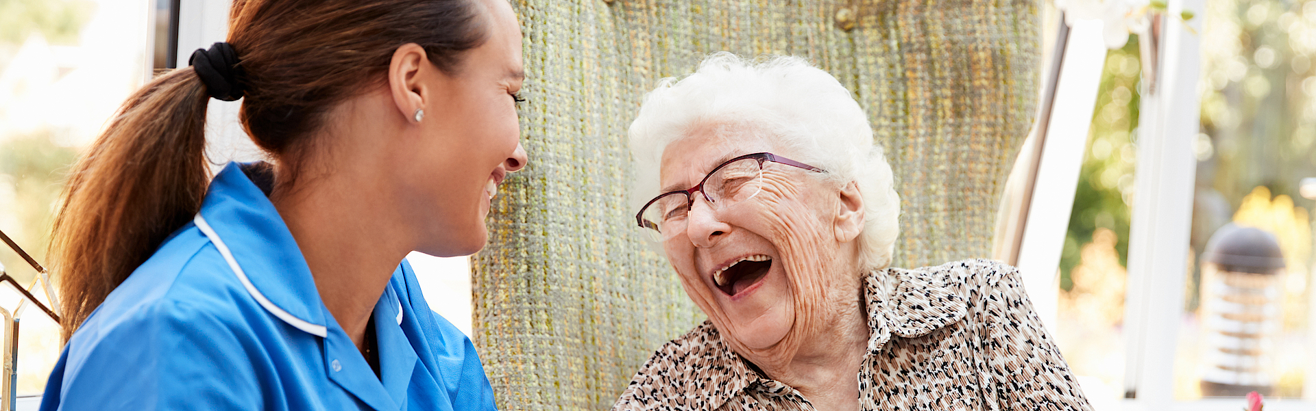 Eine Seniorin sitzt im Rollstuhl. Neben ihr eine Pflegerin im blauen Oberteil. Sie lachen beide herzlich. Ihnen geht es gut. Sie sind zusammen draußen in der Natur.