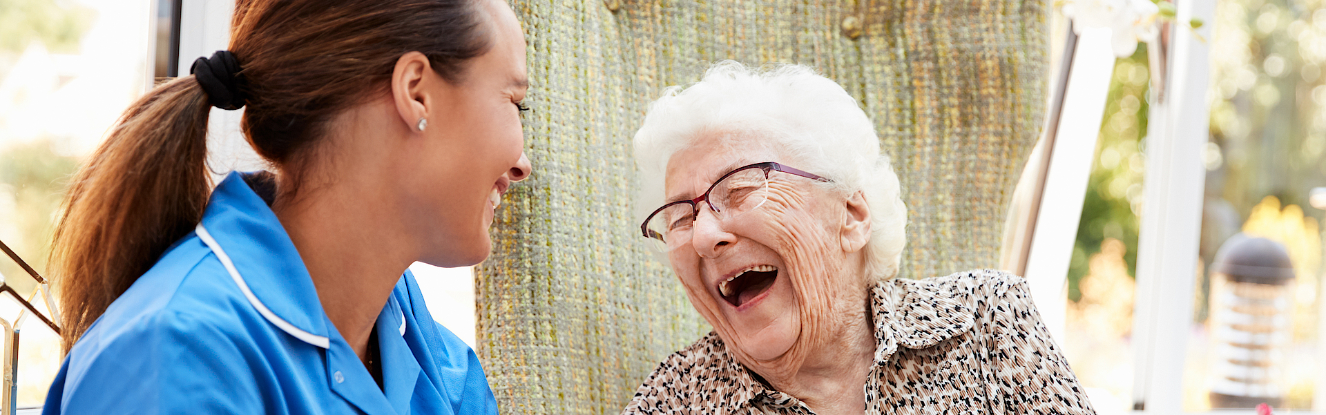 Eine Seniorin sitzt im Rollstuhl. Neben ihr eine Pflegerin im blauen Oberteil. Sie lachen beide herzlich. Ihnen geht es gut. Sie sind zusammen draußen in der Natur.