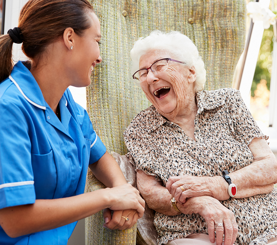 Eine Seniorin sitzt im Rollstuhl. Neben ihr eine Pflegerin im blauen Oberteil. Sie lachen beide herzlich. Ihnen geht es gut. Sie sind zusammen draußen in der Natur.