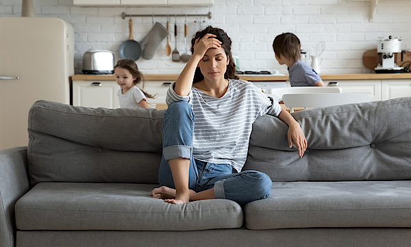 Eine Frau sitzt auf dem Sofa. Sie hat eine Jeans und ein blau-weiß geringeltes Shirt an. Sie ist depremiert. Der Kopf ist in die Hände gestützt. Ihr ist alles zu viel und zum Heulen. Hinter ihr rennen ihre Kinder.