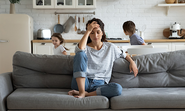 Eine Frau sitzt auf dem Sofa. Sie hat eine Jeans und ein blau-weiß geringeltes Shirt an. Sie ist depremiert. Der Kopf ist in die Hände gestützt. Ihr ist alles zu viel und zum Heulen. Hinter ihr rennen ihre Kinder.