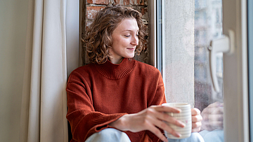 Frau im kaminroten Pullover sitzt entspannt mit einer Tasse am Fenster