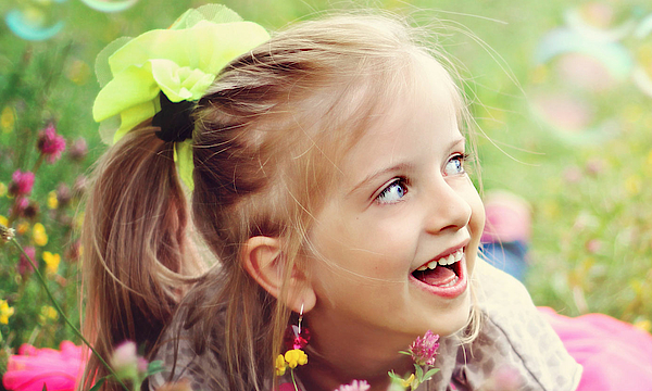 Ein kleines blondes Mädchen liegt auf einer grünen Blumenweise. Sie spielt mit Seifenblasen. Sie lacht ganz fröhlich und ausgelassen. Ihr geht es gut.