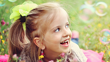 Ein kleines lachendes Mädchen mit blonden Zöpfen liegt auf einer grünen Wiese. 